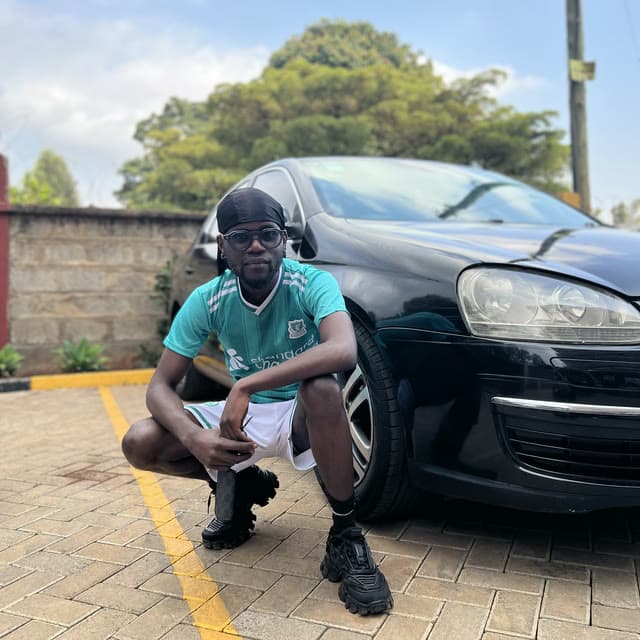 Godwill Barasa in a teal football shirt, crouching beside a black car in a paved parking bay