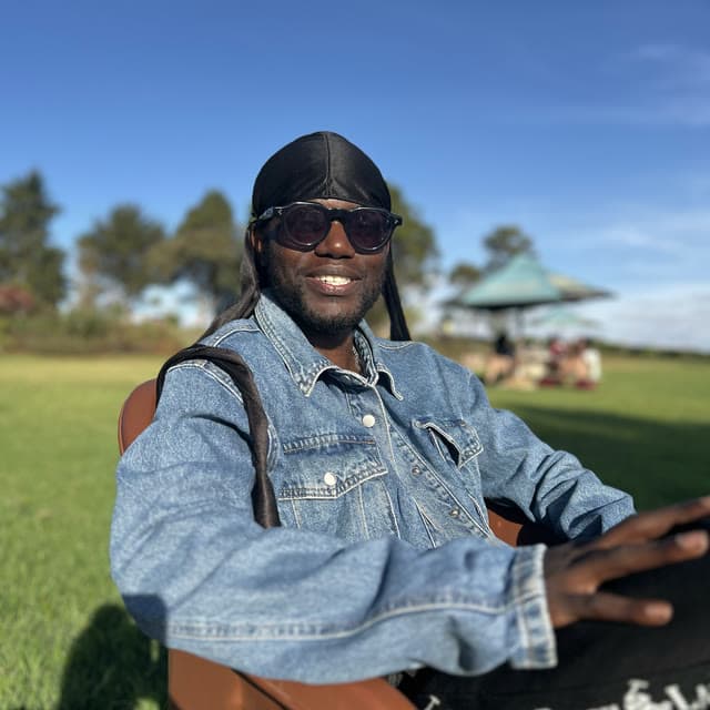 Godwill Barasa smiling in sunglasses and a denim jacket, seated outdoors on a lawn under a blue sky