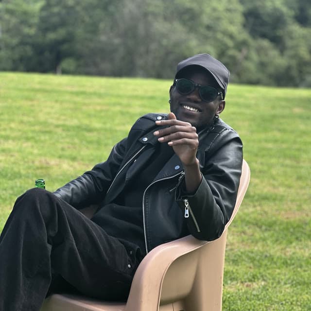 Godwill Barasa laughing in a black cap, sunglasses and leather jacket, seated on a lawn with trees behind