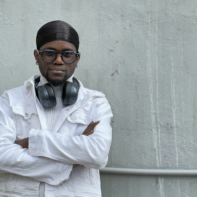 Godwill Barasa in round glasses and a white denim jacket, headphones around his neck, arms folded against a concrete wall