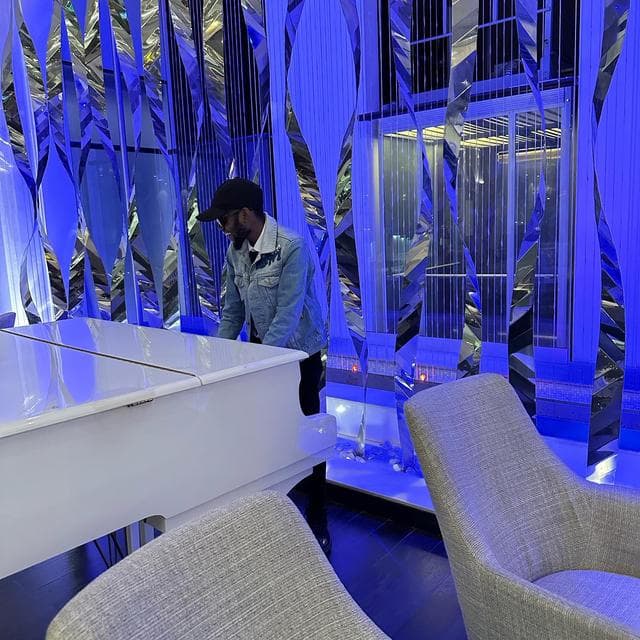 Godwill Barasa beside a white grand piano in a mirrored, blue-lit room